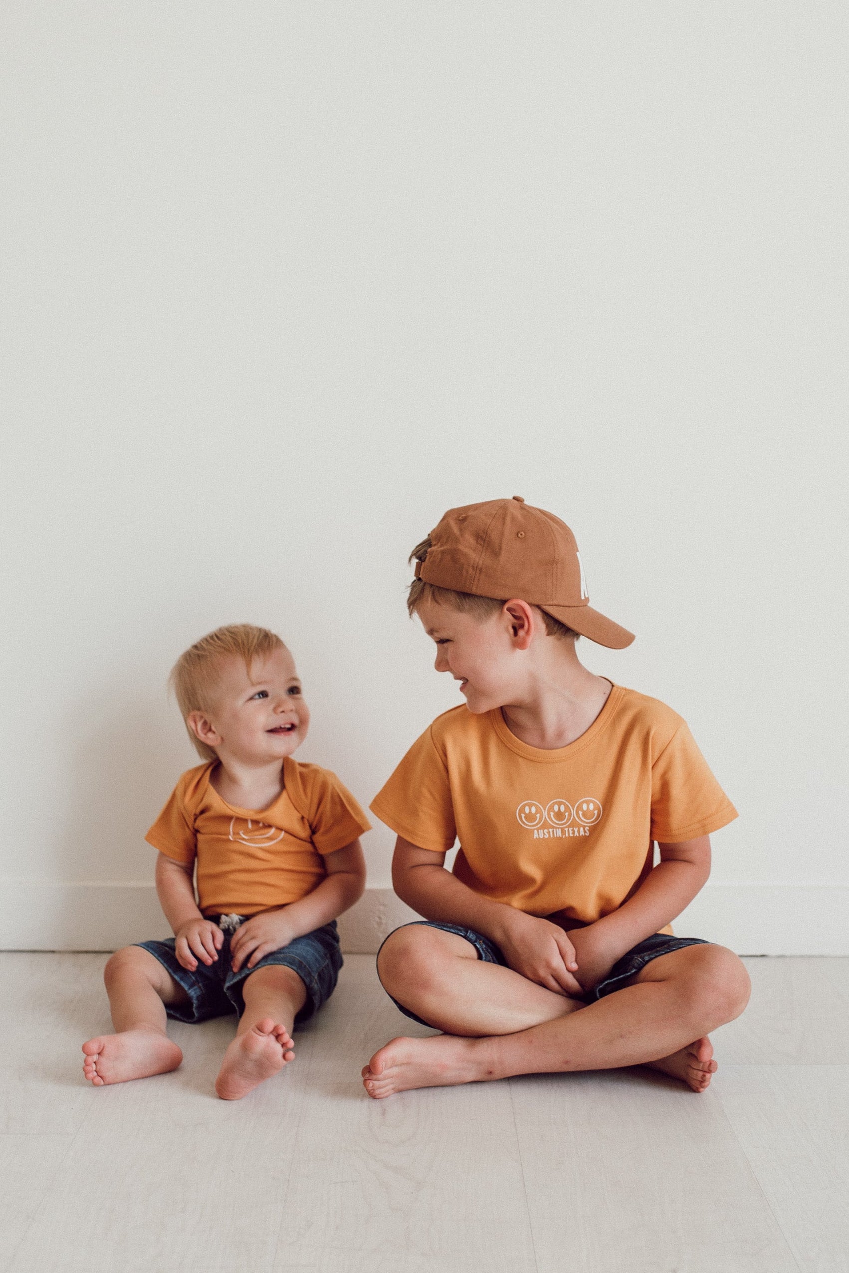 Two children sitting on a white floor wearing matching orange shirts and hats.